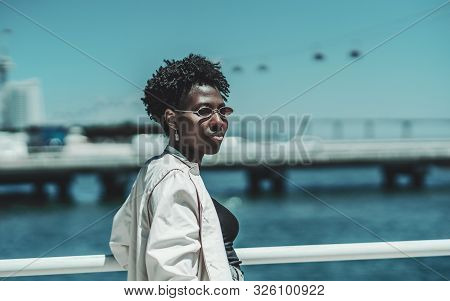 Portrait Of A Young Charming African Female In Sunglasses And White Trench Standing On The Quayside 