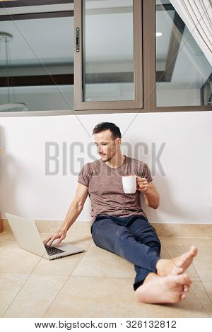 Barefoot Young Man Enjoying Big Cup Of Hot Tea And Watching Walching Film On Laptop Screen At Home