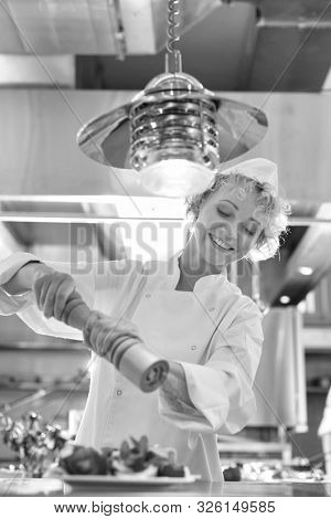 a black and white photograph of a Mature chef using peppermill on salad in plate at restaurant