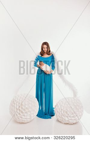 A Girl In A Blue Dress With Angel Wings Holding A Newborn Baby On A White Background. An Angel-woman