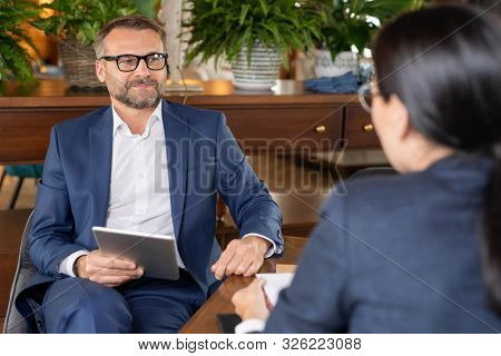 Well-dressed confident businessman with tablet sitting by table in restaurant while interacting aith colleagues at meeting