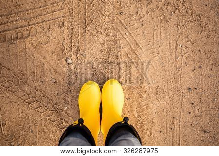 Feet In Yellow Rubber Boots Stand On A Wet Sandy Country Road
