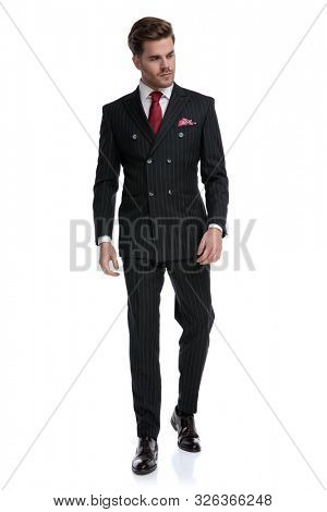 elegant young businessman wearing double breasted suit and red tie, looking to side and walking isolated on white background in studio