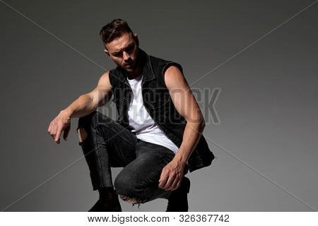 Unsure man curiously looking to the side and squatting while wearing black jeans and jeans vest on gray studio background