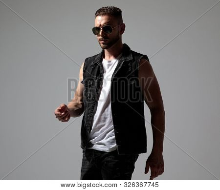 Determined man walking while wearing sunglasses and a black jeans vest on gray studio background