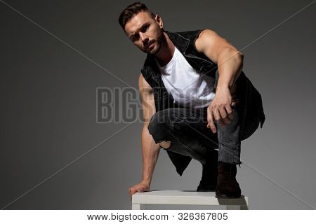 Tough casual man squatting and leaning on a chair while wearing a black jeans vest and jeans on gray studio background