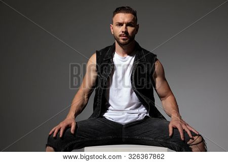 Handsome man sitting with his legs apart while wearing a black jeans vest on gray studio background
