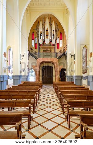 Novelda, Spain- October 3, 2019: Aisle Between Wooden Pews Leading To Pipe Organ Musical Instrument 