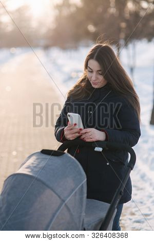 Mom Walk With Baby In Pram. Winter Park. Happy Woman With Her Baby. Mom Using Phone In Winter