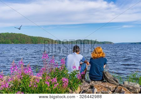 Tourists Dark-haired Man And Redhead Young Woman Sitting On Stones On Shore Of Lake Bay In Sunny Sum