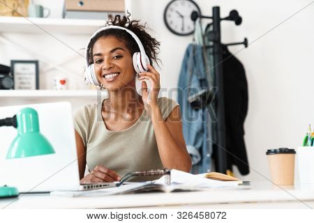 Image of a pretty happy positive optimistic young african woman sit in office using laptop computer indoors listening music with headphones.