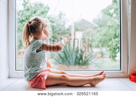 A Gorgeous Blonde Little Girl With Ponytale Staring Out Of The Window On A Wet, Cold Rainy Summer Da