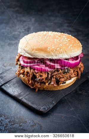 Traditional barbecue pulled beef burger offered with Carolina BBQ sauce, gherkin and onion rings as closeup on a burnt wooden board 