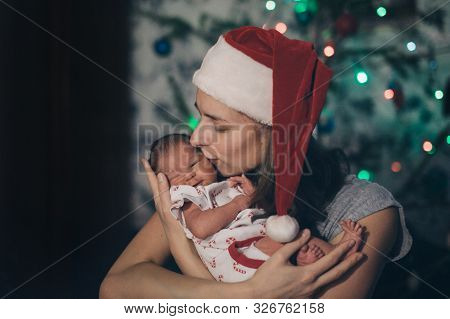 A Woman In A Christmas Hat Holding A Newborn Baby On The Background Of A Christmas Tree In Garlands.