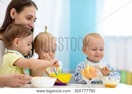 Group Of Nursery Babies Eating Healthy Food With Babysitter Help. Lunch Break In Creche. Time To Eat