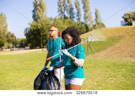 Volunteers Cleaning Park Area From Rubbish. Young Man And Woman Walking On Grass, Carrying Rakes And