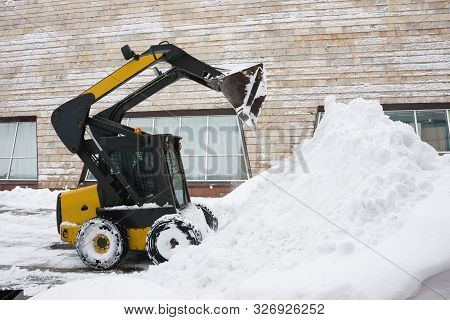 Minitractor Shovels Snow In A Pile. The Background Of The Image Is The Wall Of The Building.