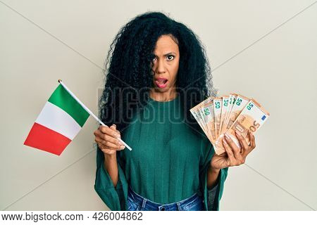 Middle age african american woman holding italy flag and euros banknotes in shock face, looking skeptical and sarcastic, surprised with open mouth 