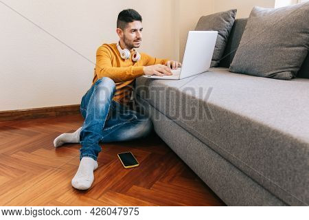 Young Adult Man 30s Doing Remote Work From Inside His Home, Sitting On The Wooden Floor Huddled On T