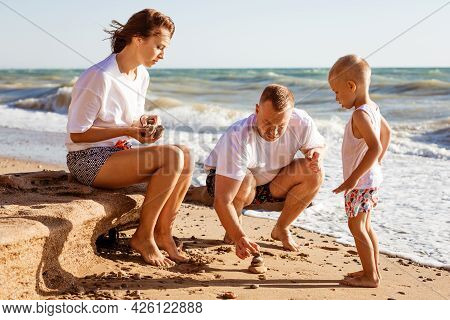 Young Family Is Painting On The Sand. Happy Parents With Their Little Son By The Sea On A Sunny Summ