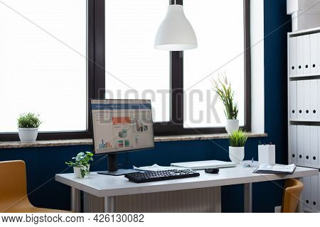 Flowers Surrounding Computer With Financial Statistics And Charts In Empty Office. Stylish Space In 