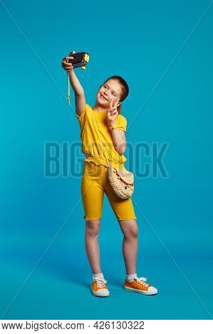 Vertical Full Body Shot Of Little Positive Girl, Takes Selfie With Camera, Makes Peace Or Victory Si