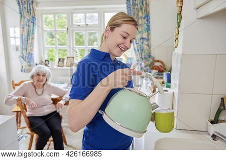 Female Home Help Making Cup Of Tea In Kitchen Whilst Chatting With Senior Woman