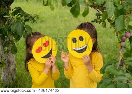 Two Children In The Park Are Standing With Their Faces Covered With Happy Emoticons And Showing Hand