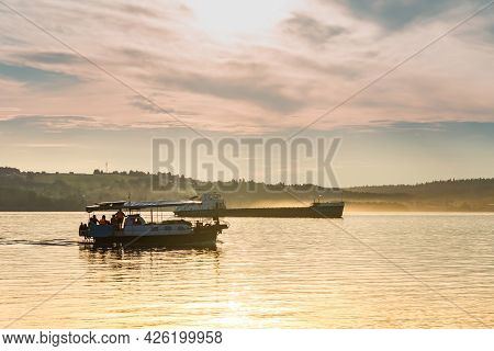 Passenger Boat And A Cargo Barge Float Down A Wide River In The Evening Light