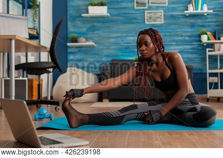 Sporty Fit Black Woman Doing Stretching Flexibility Exercise Reaching Toes Sitting On Fitness Mat In