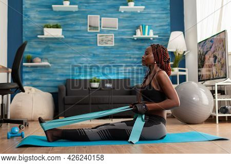 Fit Strong Afro Woman Training Home Living Room Using Resistance Band, Sitting On Fitness Mat Pullin