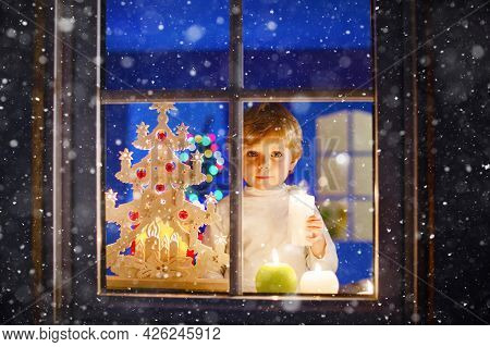 Beautiful Little Kid Boy, Standing By Window At Christmas Time And Holding Candle. Child Waiting For