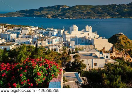 Picturesque scenic view of Greek town Plaka on Milos island over red geranium flowers and Orthodox greek church. Plaka village, Milos island, Greece