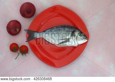 Fresh Fish On A Pink Wooden Board. Raw Uncooked Sea Bream On Red Plate. With Onions And Tomatos.