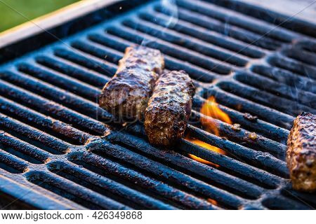 Preparing Meat Rolls Called Mici Or Mititei On Barbecue. Close Up Of Grill With Burning Fire With Fl