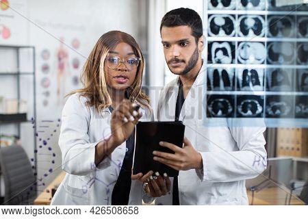 Two Multiracial Doctors Using Tablet Pc While Examining Tomography Of Patient During Consilium. Afri