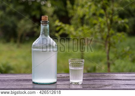 Big Bottle And Glass Of Moonshine Or Vodka On Wooden Table In Sunny Summer Day In Garden, Close Up. 