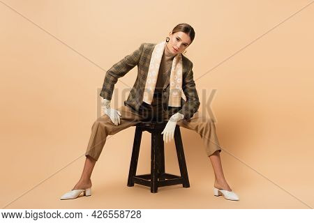 Stylish Woman In Blazer And Pants Looking At Camera While Sitting On Wooden Stool On Beige