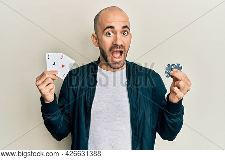 Young hispanic man playing poker holding casino cards and chips celebrating crazy and amazed for success with open eyes screaming excited. 