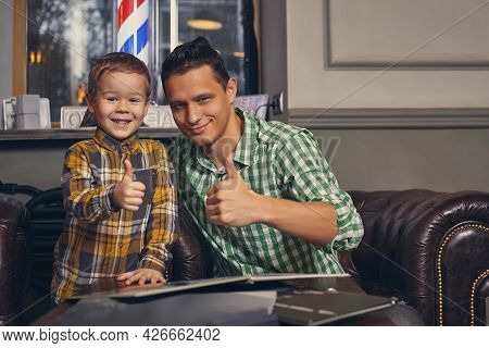 Young Father And His Stylish Little Son In The Barbershop In The Waiting Room.