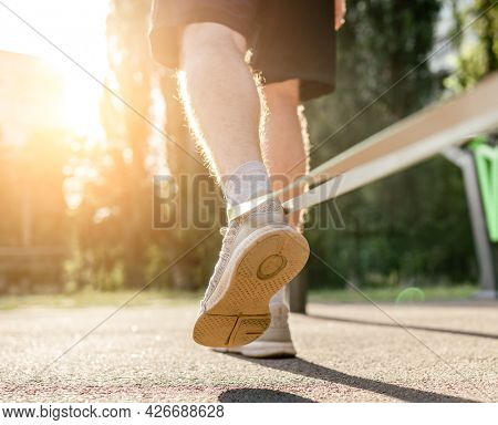 Closeup back view of legs feets of man guy training with elastic rubber band at the stadium outdoors. Adult male person during workout with additional sport equipmant with sunlight