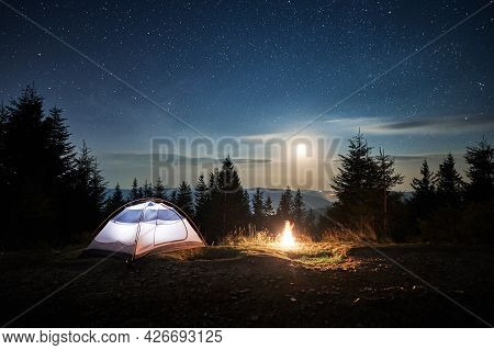 White Tourist Tent Locating On Mountain Hill Near Burning Campfire At Forest. Moonlight In The Magic