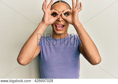 Hispanic man wearing make up and long hair wearing casual t shirt doing ok gesture like binoculars sticking tongue out, eyes looking through fingers. crazy expression. 