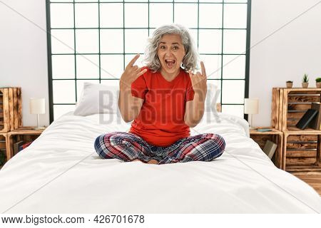Middle age woman with grey hair sitting on the bed at home shouting with crazy expression doing rock symbol with hands up. music star. heavy concept. 
