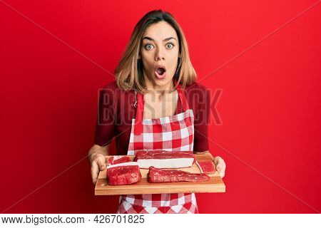 Young caucasian blonde woman holding board with raw meat afraid and shocked with surprise and amazed expression, fear and excited face. 