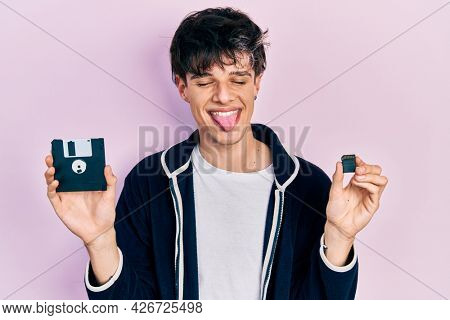 Handsome hipster young man holding floppy disk and sdxc card sticking tongue out happy with funny expression. 