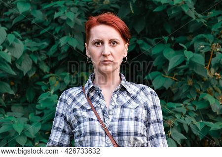 Woman In A Checkered Shirt Stands On A Background Of Green Leaves Of Bush. Serious, Worried Expressi