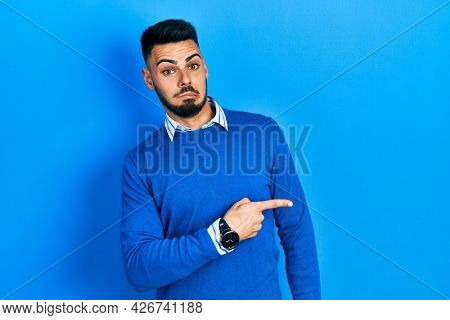 Young hispanic man with beard wearing casual blue sweater pointing aside worried and nervous with forefinger, concerned and surprised expression 