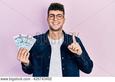 Young hispanic man holding peruvian sol banknotes smiling with an idea or question pointing finger with happy face, number one 
