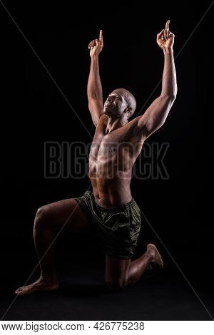 Black Adult In Shorts Kneeling With Arms Extended Upward On Black Background.
